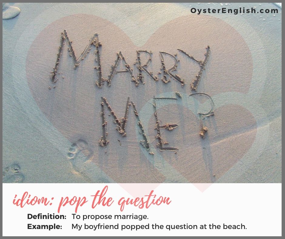 The words "Marry Me" written in the beach sand within a red heart design. Idiom "pop the question": to propose marriage; Example: My boyfriend popped the question at the beach.