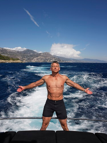 A man is standing at the rear of the boat with his arms stretched out enjoying the breeze from a boat. The wake is visible behind the boat as a stream of white water and waves.